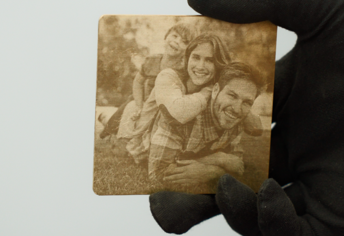 Рука в перчатке держит металлический квадрат с гравировкой семейного фото: родители и ребёнок улыбаются на природе