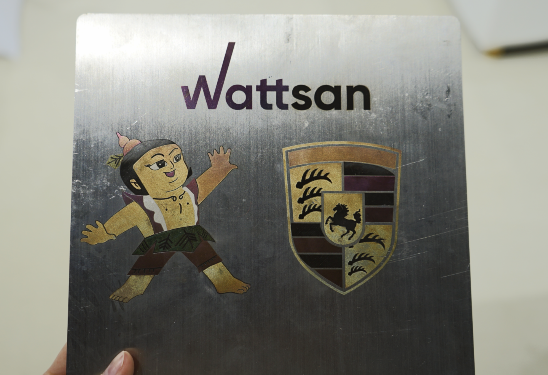 Рука держит металлическую пластину с "Wattsan", мультяшным персонажем в шляпе и логотипом Porsche